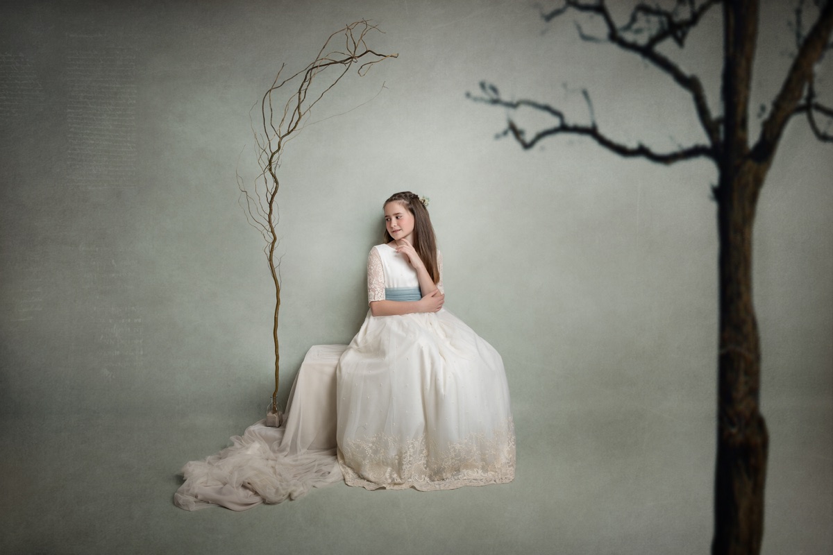 Retrato de una niña de Comunión junto a un árbol en una escena de estudio.