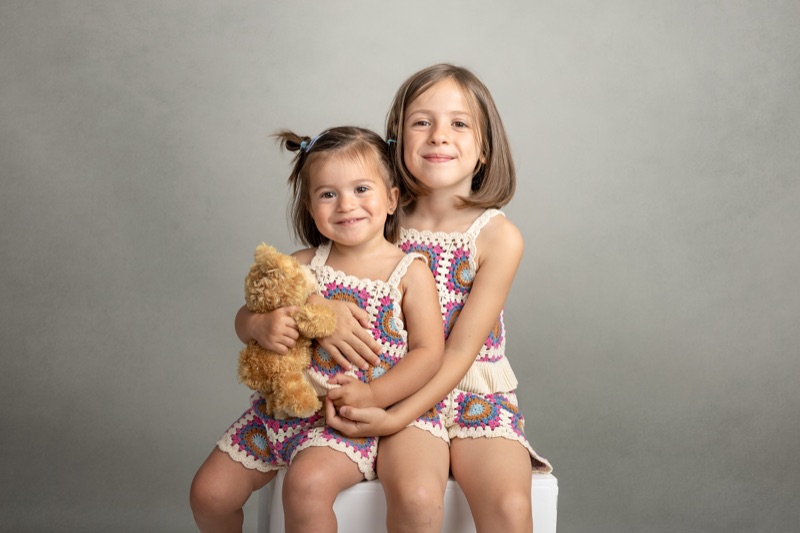 Dos hermanas sentadas juntas con vestidos de ganchillo y un peluche durante una sesión infantil