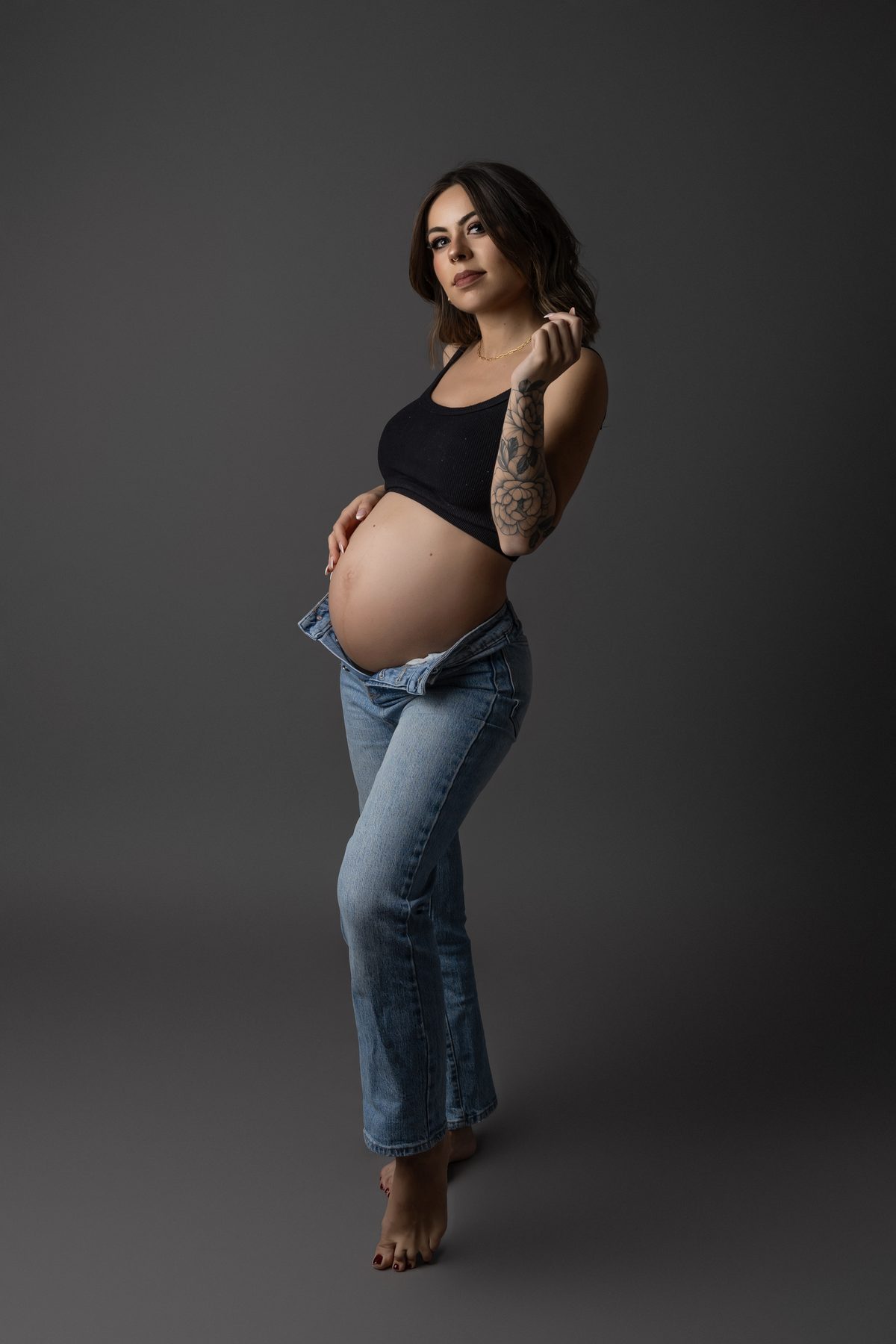 Retrato de cuerpo entero de una futura mamá con vaqueros y top negro sobre un fondo oscuro.