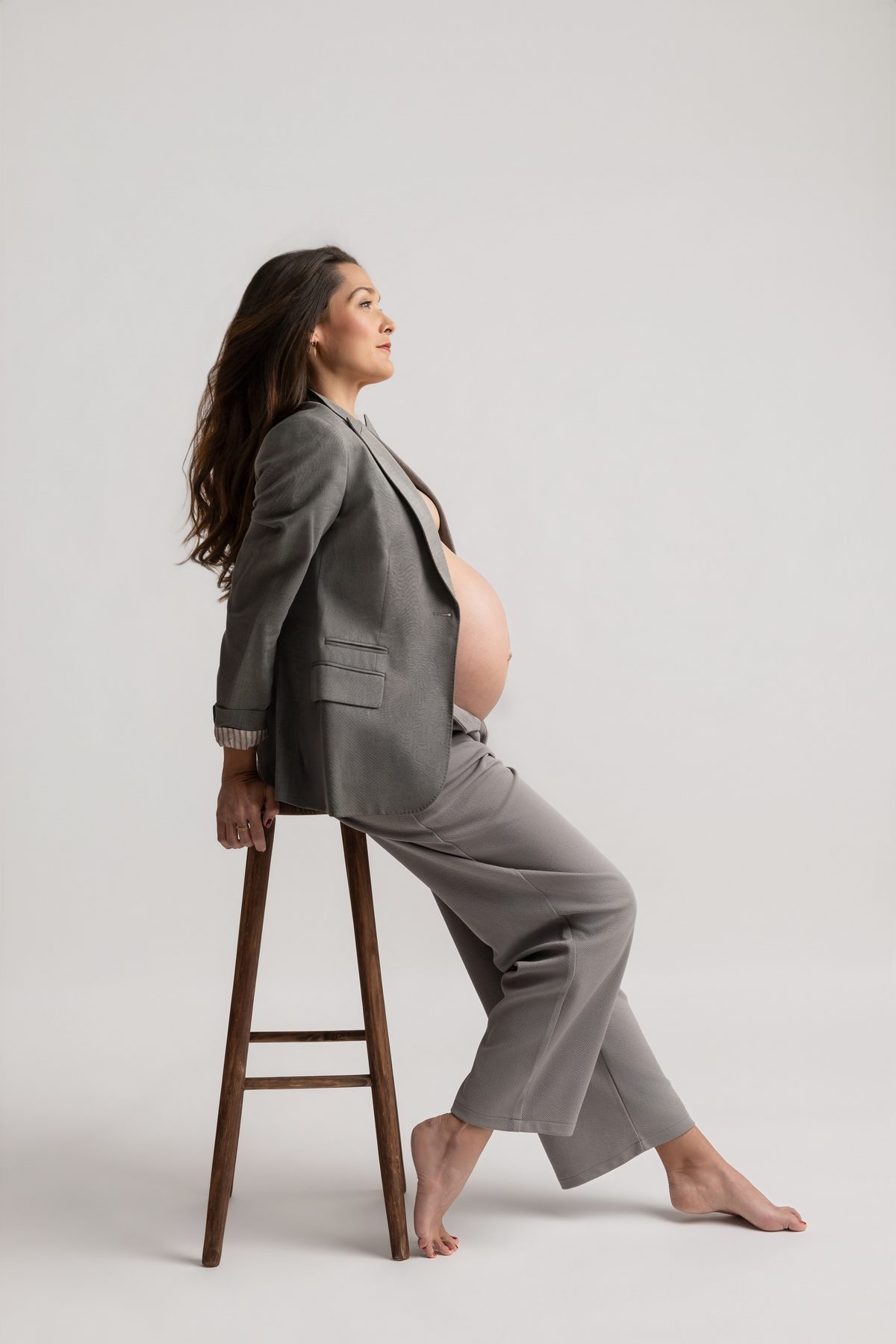 Retrato de maternidad con traje gris, sentada en un taburete sobre un fondo claro.