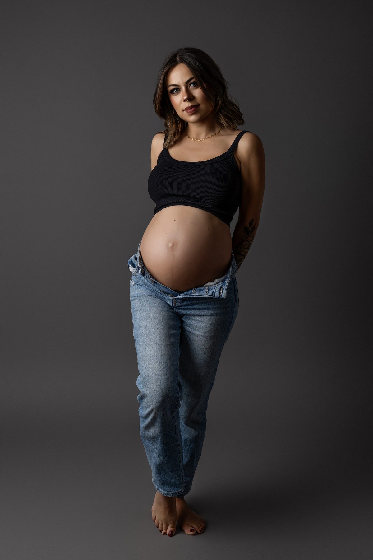 Retrato de cuerpo entero de maternidad con vaqueros y top negro, sobre un fondo oscuro.