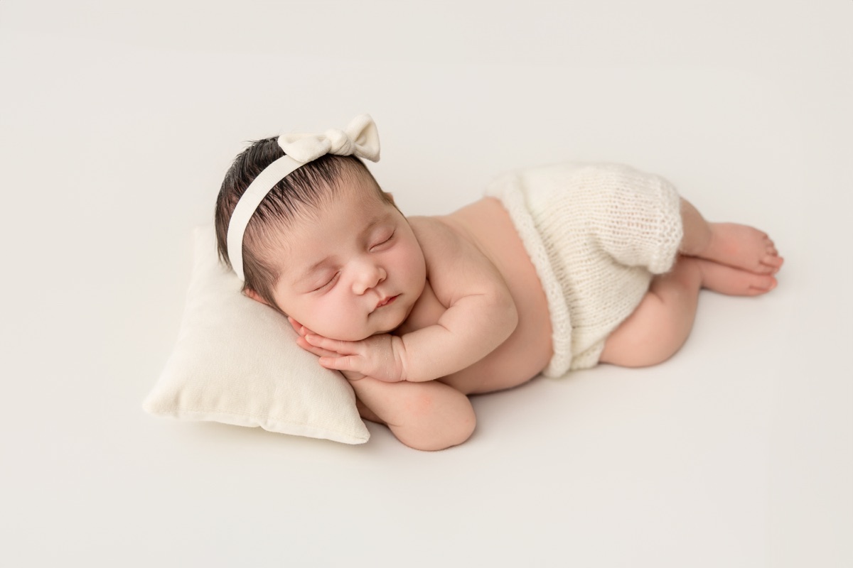 Bebé recién nacida dormida sobre un cojín blanco con un lazo de punto durante una sesión newborn en Villanueva de la Cañada