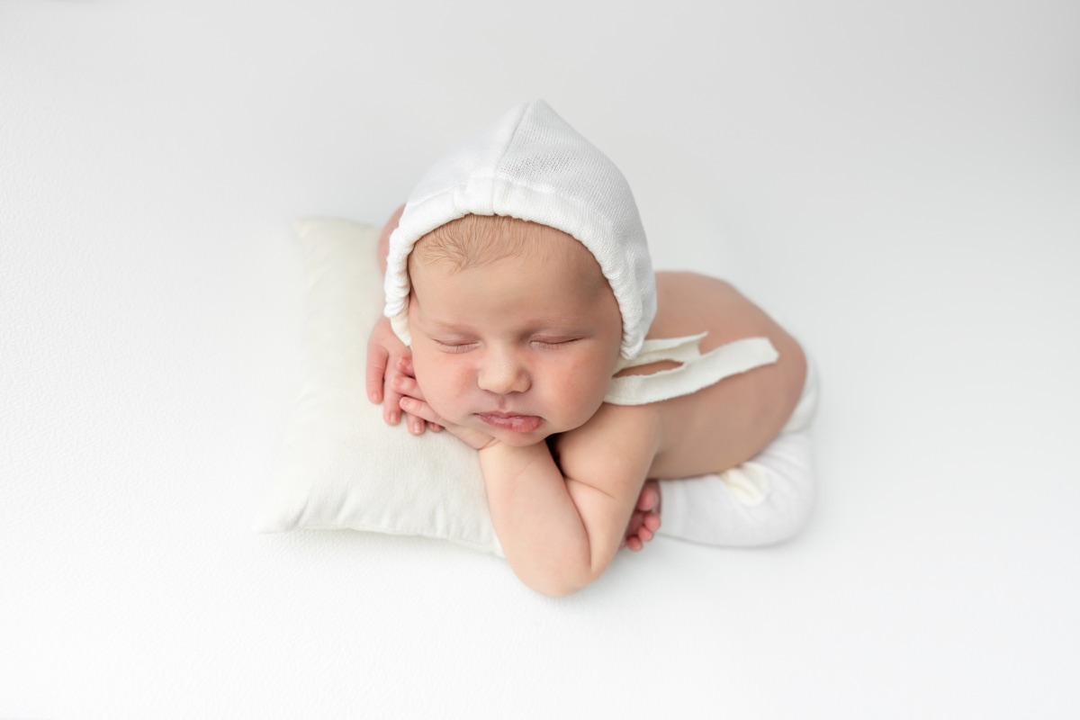 Bebé recién nacido dormido boca abajo con gorrito blanco sobre un fondo claro durante una sesión de fotografía newborn en Villanueva de la Cañada