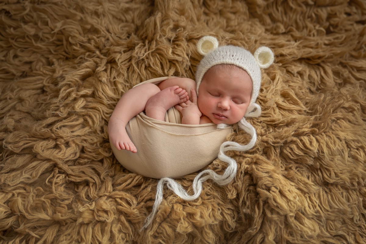 Bebé recién nacido acurrucado en un cuenco sobre una manta de textura marrón, con gorrito de punto durante una sesión de fotografía newborn en Villanueva de la Cañada
