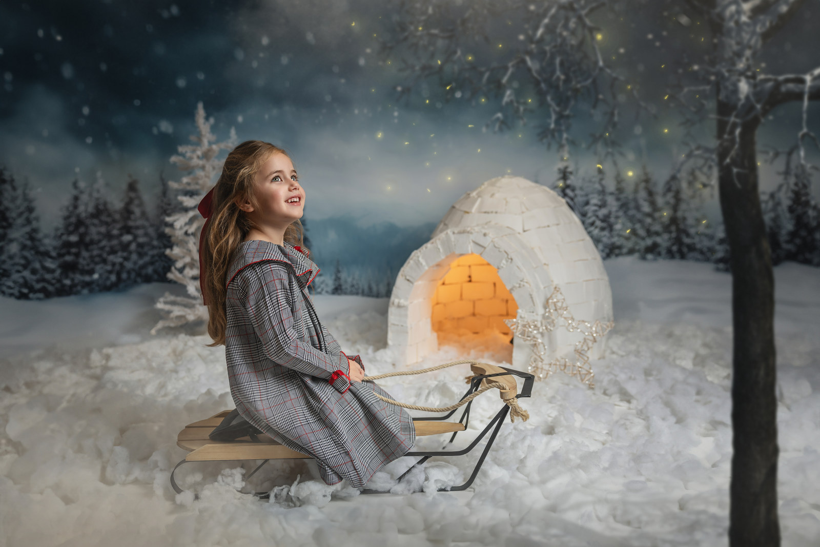 Niña con abrigo rojo junto a un trineo y un iglú en una sesión de Navidad